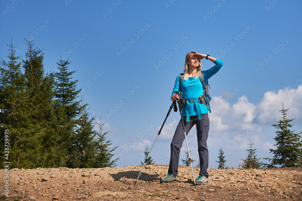Female tourist standing on dirty stone road and covering her face with hand protecting it from bright sunlight, while she watching and studying her next way in hiking. Concept of travelling, hiking.
