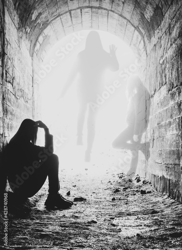 a dying young woman sits in a tunnel in bright rays of light. the soul of the girl opposite the body.