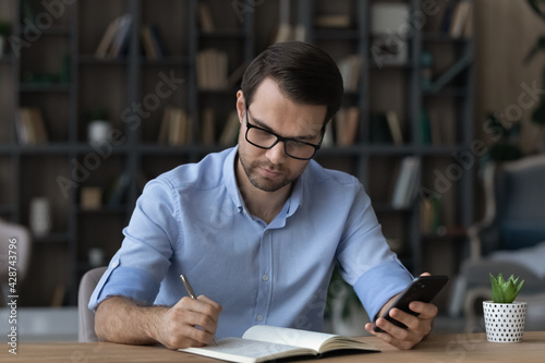 Pensive young Caucasian man sit at desk use cellphone make note or list in notebook. Thoughtful male write in notepad, engaged in time management or planning with smartphone application online.