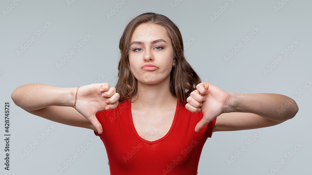 Portrait of a cute young brunette woman in red casual t shirt showing thumbs down. Gray background Portrait of a cute young brunette woman in red casual t shirt showing thumbs down. Gray background