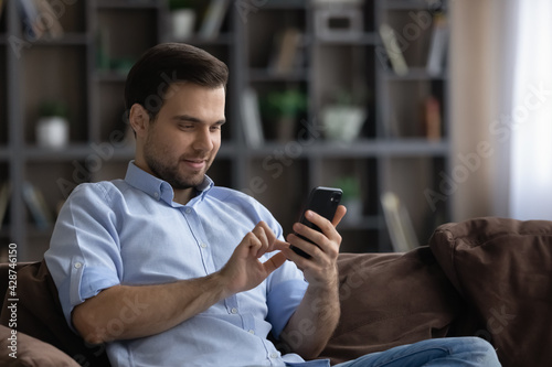 Smiling young Caucasian man relax sit on sofa at home use cellphone gadget texting messaging. Happy millennial male browse wireless Internet on smartphone device online. Communication concept.