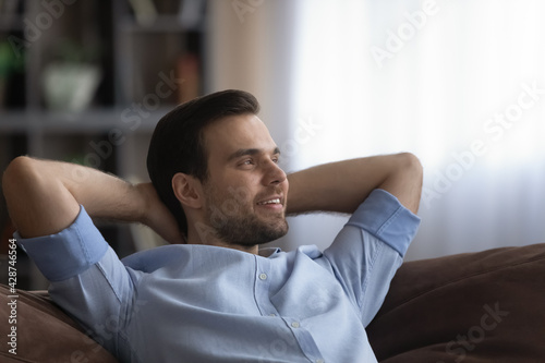 Happy Caucasian man sit rest on couch at home look in window distance dreaming thinking of new opportunities. Smiling young male make plans or decision, relax in living room. Vision concept.
