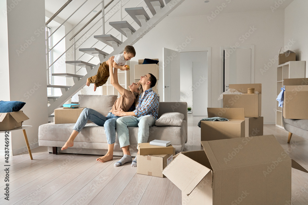 Happy young family couple playing with small toddler kid son together in new home. Parents and child boy having fun in new house living room with boxes. Moving day, mortgage, relocation concept