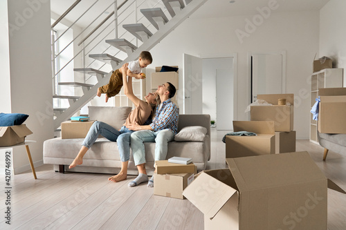 Happy young family couple playing with small toddler kid son together in new home. Parents and child boy having fun in new house living room with boxes. Moving day, mortgage, relocation concept