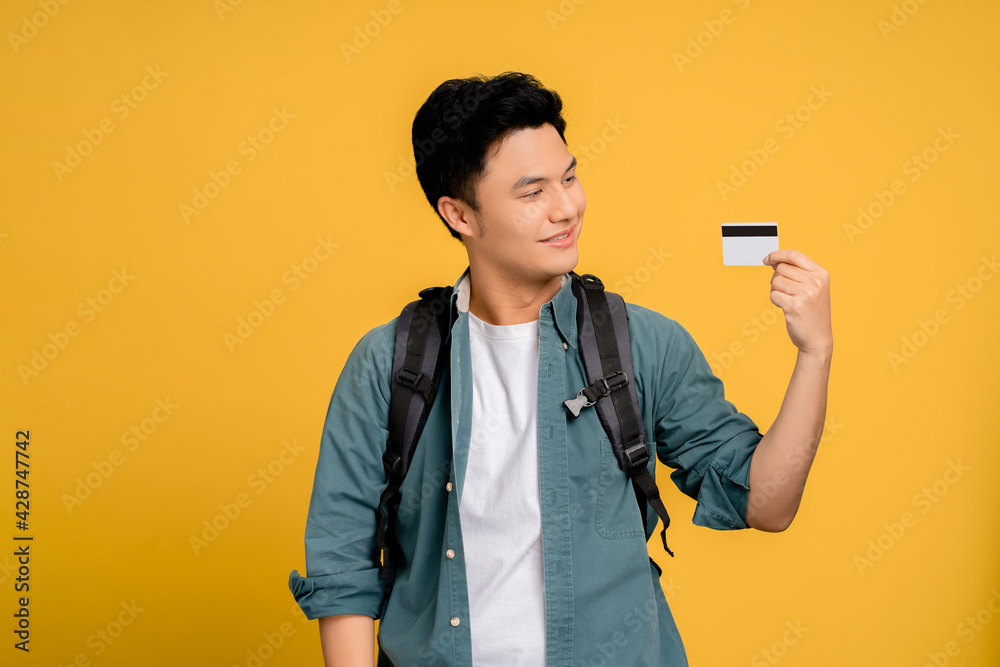 Young Asian man in casual wear holding credit card on yellow background