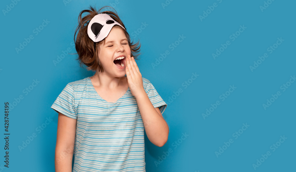 Photo of adorable sweet kid nightwear yawning stretch. Sleepy child girl isolated on blue background yawns, sleep mask on her head. Banner design. Copy space Photo of adorable sweet kid nightwear yawning stretch. Sleepy child girl isolated on blue background yawns, sleep mask on her head. Banner design. Copy space