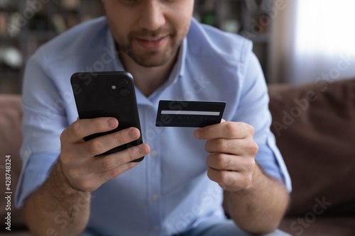 Crop close up of young Caucasian man hold cellphone shopping online with credit card at home. Millennial male client or buyer buy on internet on smartphone with secure banking service system.