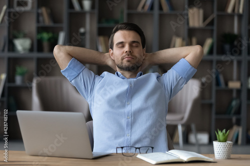 Calm young Caucasian businessman sit lean in chair at workplace in home office breathe fresh condition air. Happy man relax rest at desk take nap or sleep, relieve negative emotions. Peace concept.