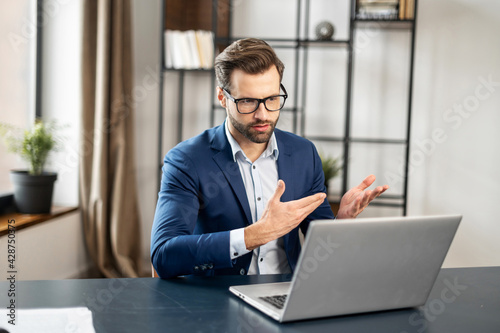 Slavic stylish man talking at laptop by video call, focused young businessman in eyeglasses involved in online video call negotiations meeting with partners colleagues or studying distantly at home