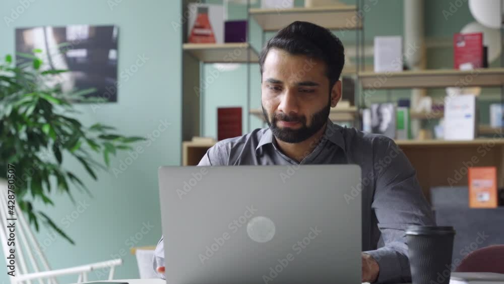 Young bearded indian businessman working on laptop in modern office lobby space. Indian executive using computer remote studying, watching online webinar, virtual training from homeoffice.