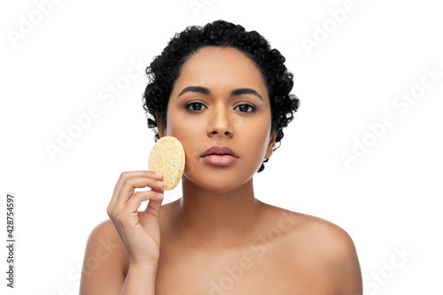 beauty  people and skincare concept - young african american woman with bare shoulders cleaning face with exfoliating sponge over white background