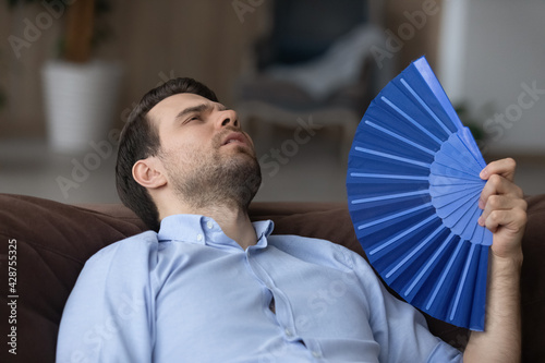 Unhappy tired young Caucasian man relax on sofa at home wave with hand fan suffer from heatstroke. Exhausted millennial male use waver breathe fresh air, struggle lack conditioner indoors.
