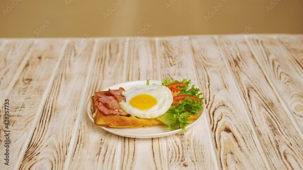 Morning breakfast belgian waffle with eggs, bacon, tomatoes and lettuce salad. Healthy breakfast in the morning, slow motion view, close up. On white plate over wooden table. Morning breakfast belgian waffle with eggs, bacon, tomatoes and lettuce salad. Healthy breakfast in the morning, slow motion view, close up. On white plate over wooden table.