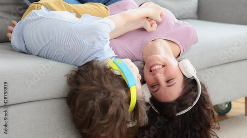 Smiling happy mother and kid son lying upside down on couch at home, laughing, embracing, hearing songs in earphones. Excited child and mom wear headphones enjoy listening to music together.