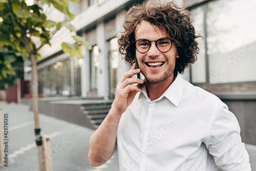 Outdoor image of a young man with curly hair smiling while talking with his friend on a mobile phone. A happy curly male wearing a white shirt has a joyful expression during speaking on a cellphone.