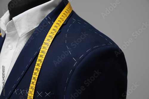 Semi-ready jacket with tailor's measuring tape on mannequin against grey background, closeup photo