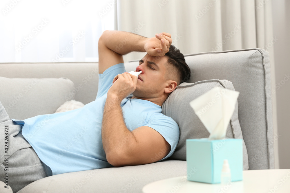 Ill man using nasal spray on sofa at home Ill man using nasal spray on sofa at home