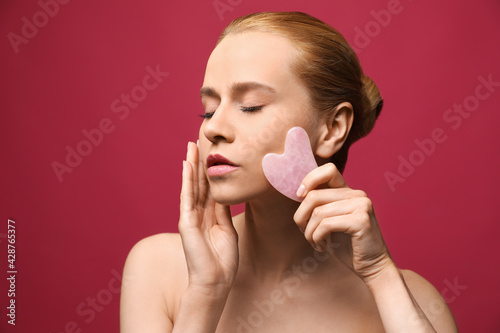 Beautiful young woman doing facial massage with gua sha tool on pink background