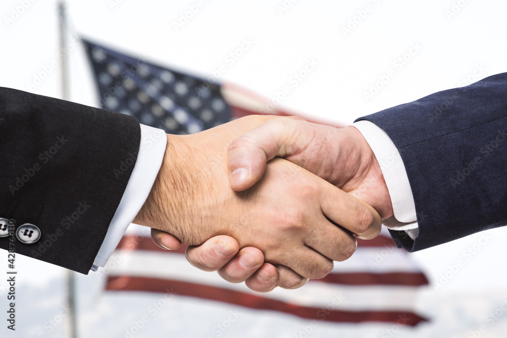 Two businessmen shake hands on the background of USA flag, partnership concept, close up Two businessmen shake hands on the background of USA flag, partnership concept, close up