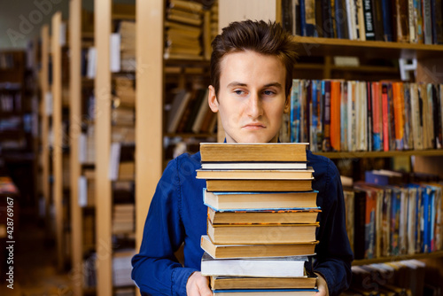 Male student with negative and bored mimic on his face holding a big stack of books in the library. Concept of hard education, learning