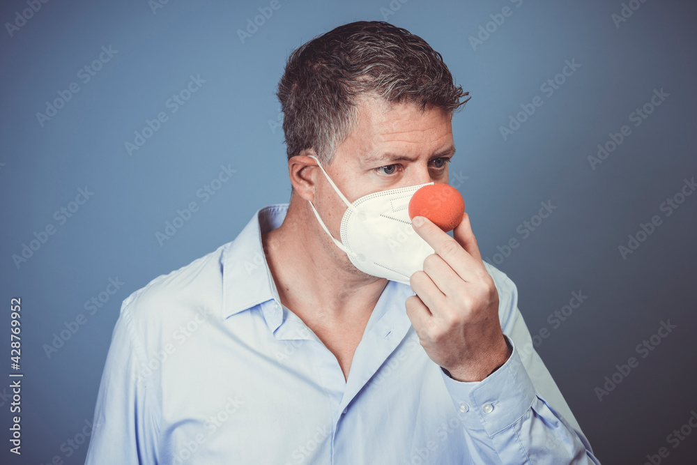Middle aged man with nose mouth protection and red clown nose and blue shirt in front of blue background Middle aged man with nose mouth protection and red clown nose and blue shirt in front of blue background