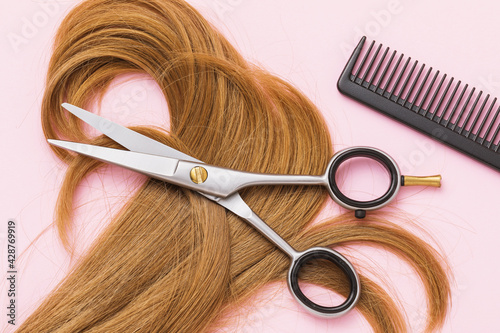 Scissors, a comb and a lock of curly blond female childrens hair on a pink background, top view