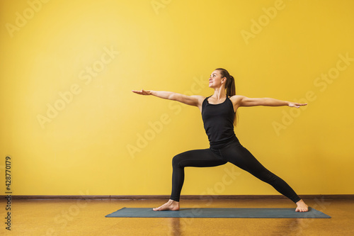 Attractive woman in black sportswear practicing yoga performs Virabhadrasana exercise, warrior pose 2, against a yellow wall with copy space