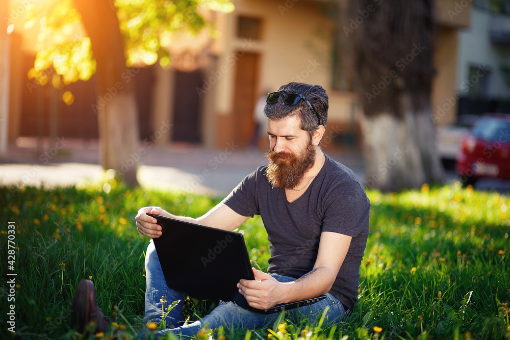 © endrews21 - A man with a long beard womanizer and dark hair with gray hair in a shirt and jeans sits on green grass in a park with a laptop and watches funny videos © endrews21 - A man with a long beard womanizer and dark hair with gray hair in a shirt and jeans sits on green grass in a park with a laptop and watches funny videos