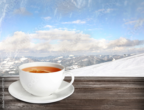 Cup of hot tea on wooden table and beautiful winter landscape on background