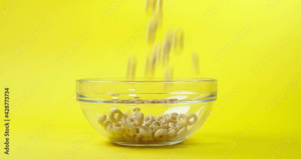 Cornflakes in the form of rings fall into a transparent plate on an isolated yellow background in slow motion in 4K. healthy breakfast of cereal on a yellow background. Cornflakes in the form of rings fall into a transparent plate on an isolated yellow background in slow motion in 4K. healthy breakfast of cereal on a yellow background.