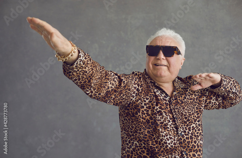 Retired senior man vibing to hype pop music. Portrait of funny rich white haired old grandpa in cool glasses and leopard patterned party shirt flexing, dancing and having fun on gray studio background photo