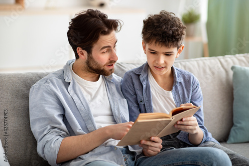 Family pastime. Father and teen boy reading book together, resting on comfy sofa in living room