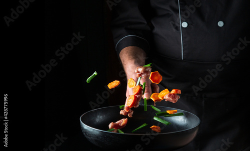 The hand of a professional chef tosses pieces of vegetables and meat on a frying pan on a black background. Restaurant cooking concept. Free advertising space