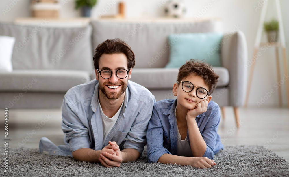 Portrait of cheerful dad and son wearing eyeglasses and smiling to camera while lying on floor carpet - obrazy, fototapety, plakaty 