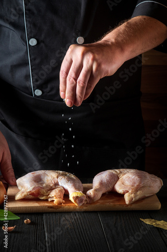 The chef adds salt to the raw chicken legs. Preparation for grilling with chicken leg in the kitchen of a restaurant or cafe on table with vegetables and spices