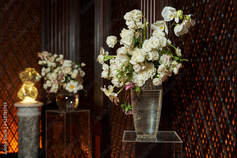 two white bouquet of orchids, roses and other flowers near a carved wooden wall on transparent stands in large transparent vases