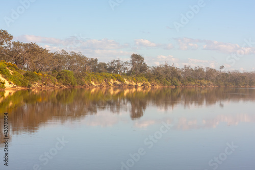 Reflections of clouds, blue sky and riverside trees and vegetation in the calm waters of a river in east coast Australia highlighted by a golden haze created by a summer dust storm.