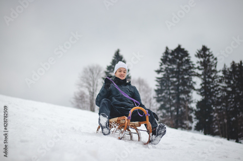 Joyful smile of a young sixteen year old boy riding a historic wooden sled on a ski slope. Dangerous driving. Enjoying the fallen snow in winter. Fooling around at a young age. Immortality.