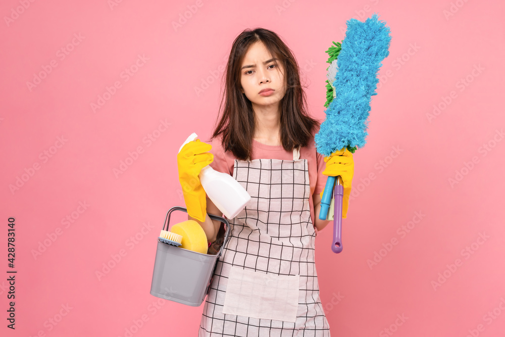 Boring young Asian woman wearing apron with yellow rubber gloves and holding bucket of cleaning supplies on pink background. Boring young Asian woman wearing apron with yellow rubber gloves and holding bucket of cleaning supplies on pink background.