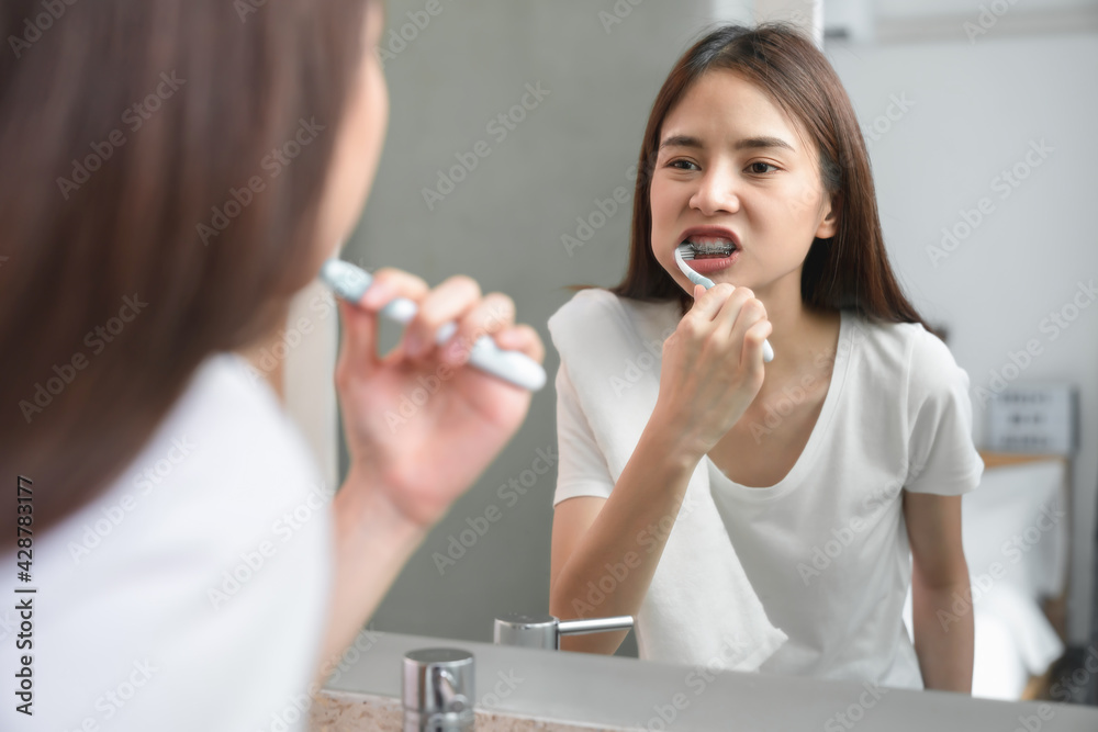 Young Asian woman wearing braces with brushing teeth and looking in the mirror, holding water glass, towel on the shoulder on bathroom, Concept oral hygiene and health care. Young Asian woman wearing braces with brushing teeth and looking in the mirror, holding water glass, towel on the shoulder on bathroom, Concept oral hygiene and health care.