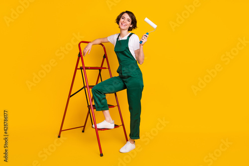 Full size photo of happy excited positive good mood woman painter with ladder hold roller isolated on yellow color background