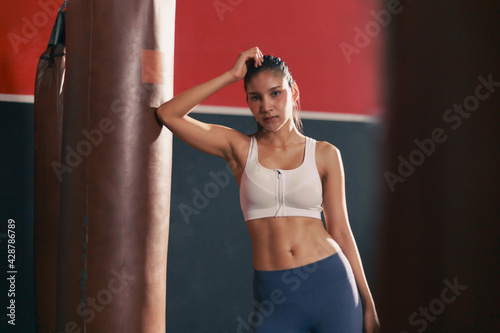 Portrait of young fit Asian woman standing by the punching bag looking at camera boxing exercise indoor at sport club fitness center © butsaya33