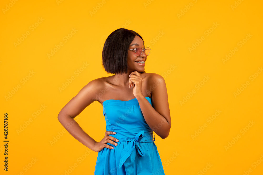 Pretty black lady in blue dress and sunglasses touching her chin and looking aside on orange studio background Pretty black lady in blue dress and sunglasses touching her chin and looking aside on orange studio background