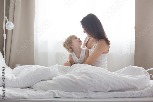 Beautiful blond child and his mother, playing at home in the morning in bed, smiling, laughing and having fun