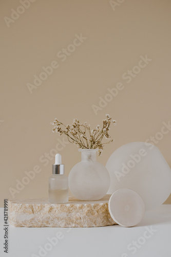 Aesthetic minimalist beauty care therapy concept. Organic serum oil cosmetics bottle on stone with flowers against neutral beige background. Body skin, face treatment product composition