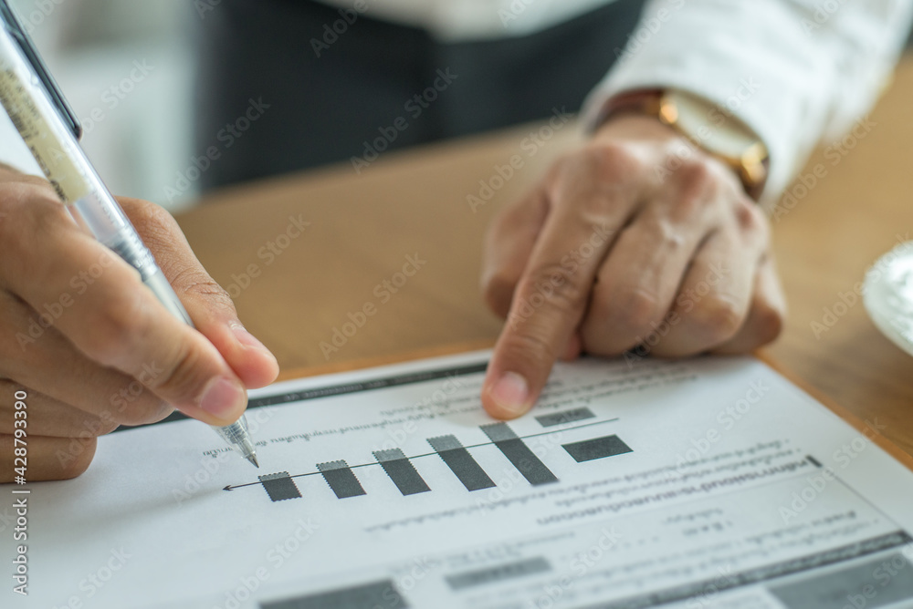 Businessman wearing white shirt analyzing income charts and graphs with hand holding pen on office desk. Close up.Business analysis and strategy concept. Businessman wearing white shirt analyzing income charts and graphs with hand holding pen on office desk. Close up.Business analysis and strategy concept.