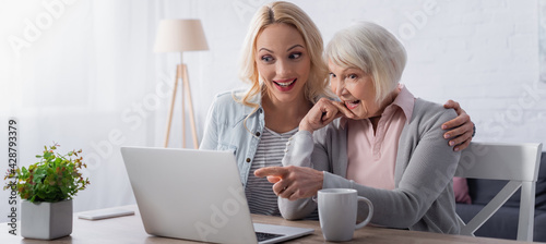 Cheerful woman pointing with finger at laptop near tea and daughter, banner