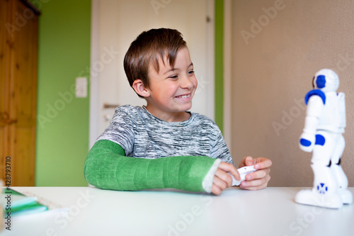 Billede på lærred cool boy with green arm cast is sitting in his room and is playing with toy robo