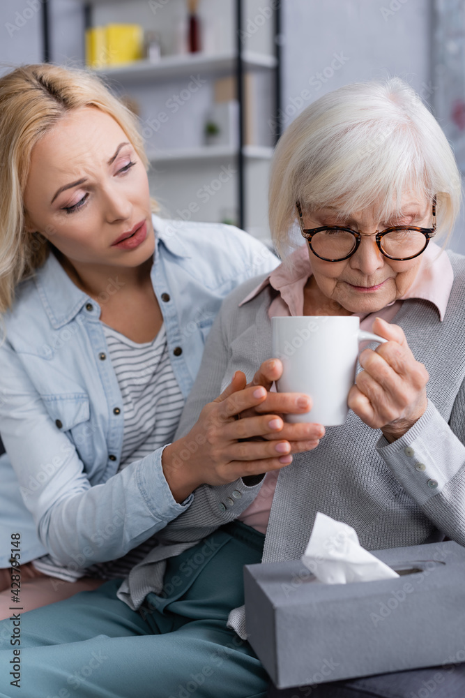 Adult woman calming senior mother with cup near napkin Adult woman calming senior mother with cup near napkin