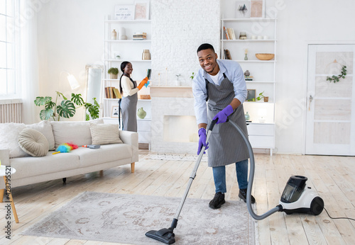 Full length of black guy vacuuming floor, his girlfriend helping him to clean living room, empty space